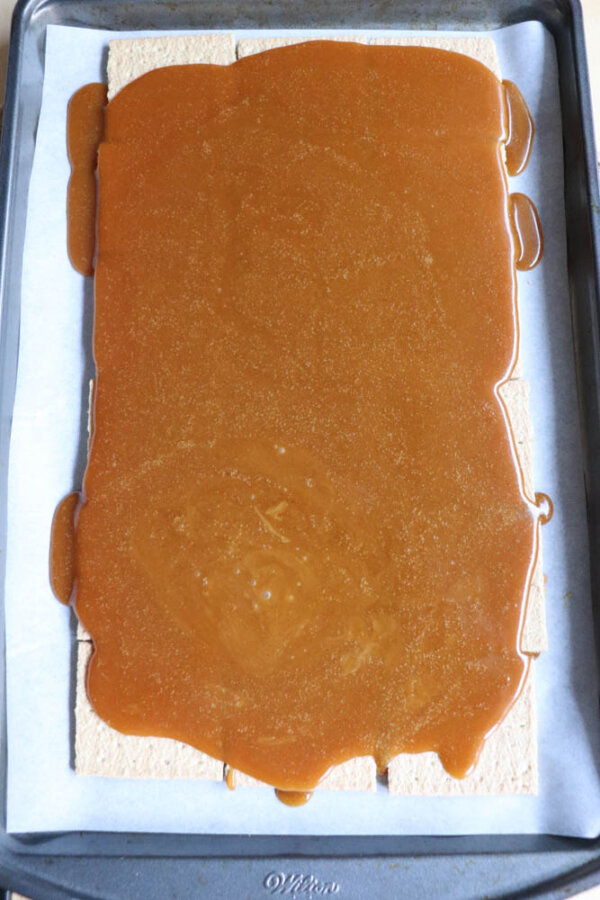 A baking tray lined with parchment paper holds a layer of graham crackers covered with a thick, even layer of caramel sauce, some of which has slightly spilled over the edges—an irresistible step in this smores crack recipe.
