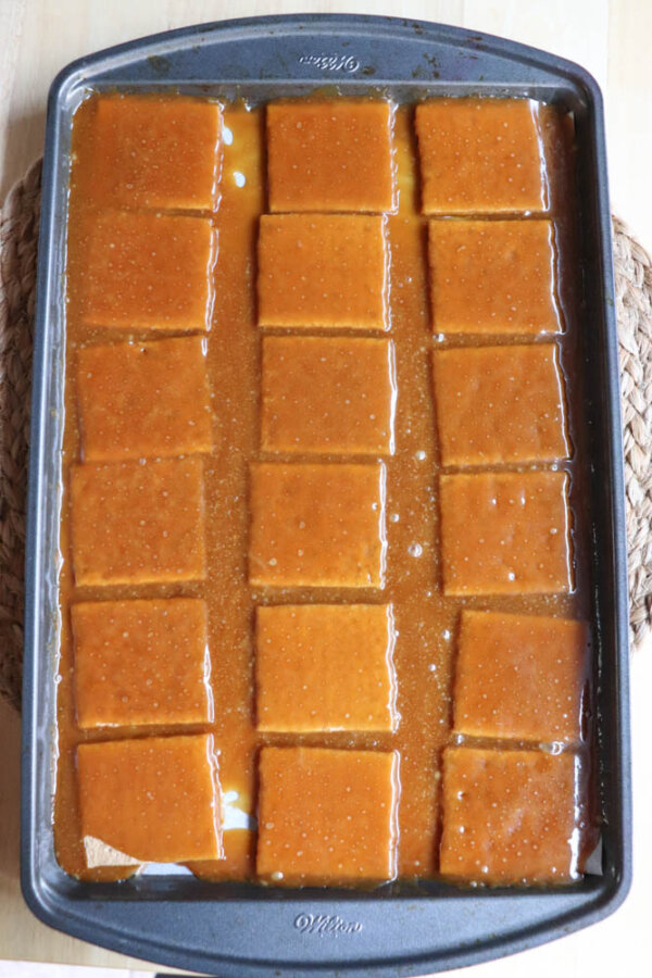 A baking pan filled with 18 neatly cut squares of caramel flan, with a shiny caramel sauce like that in a smores crack recipe covering the surface and pooling around the edges. The pan is placed on a woven mat.