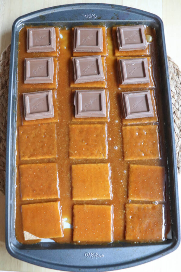 A baking tray with rows of graham crackers topped with caramel sauce; the top row features squares of milk chocolate, making this an irresistible smores crack recipe.