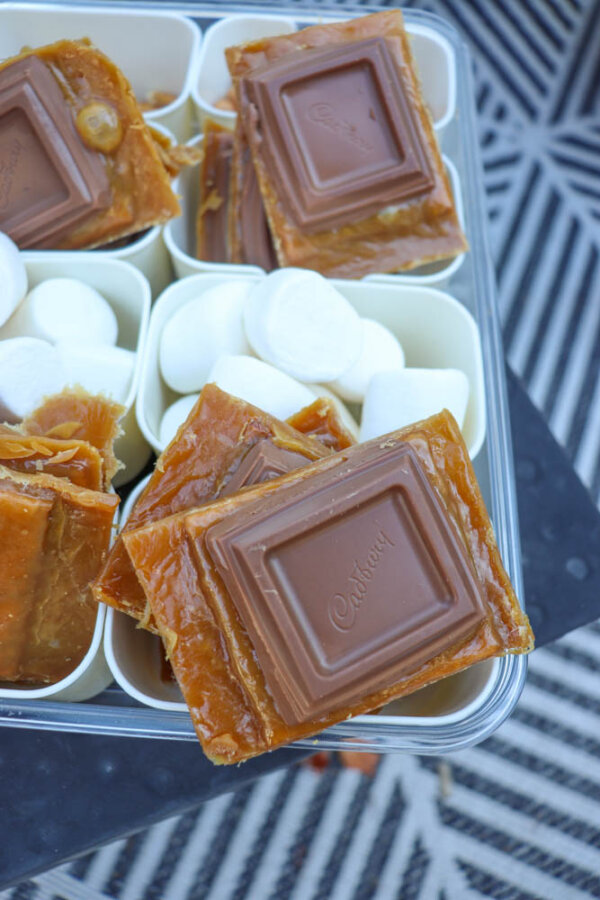 Close-up of a tray featuring pieces of caramel toffee topped with Cadbury milk chocolate squares, inspired by a smores crack recipe. Sections are filled with white marshmallows on a black-and-white patterned surface underneath.