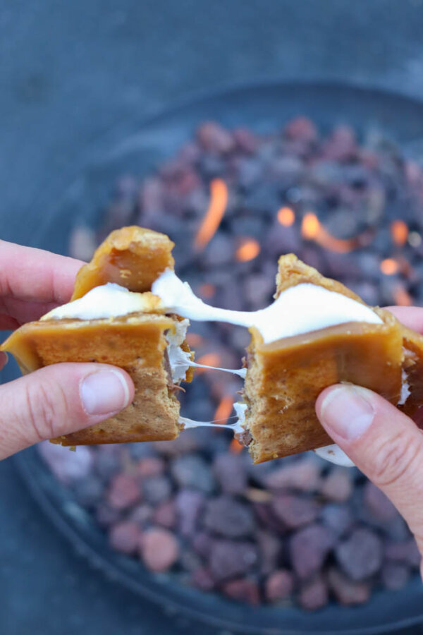 Two hands pull apart a gooey s’more—with toasted marshmallow and melty chocolate, inspired by a classic s’mores crack recipe—between graham crackers, with a fire pit and glowing coals blurred in the background.