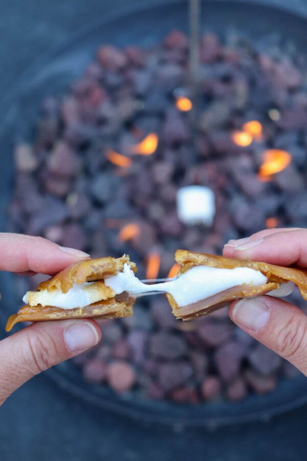 Two hands pull apart a gooey smores crack recipe, with toasted marshmallow and caramel between two cookies, held in front of a fire pit with flames and a roasting marshmallow in the background.