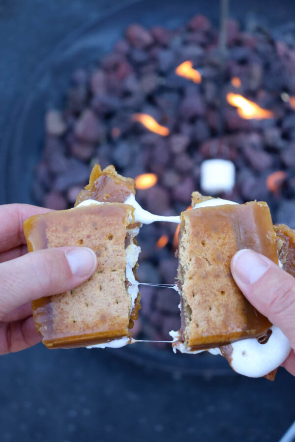 Two hands pull apart a gooey caramel smore with melted marshmallow, reminiscent of the popular smores crack recipe, with a campfire and roasting marshmallow blurred in the background.