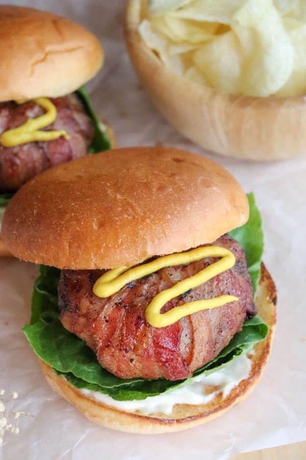 A bacon-wrapped burger made from our signature bacon wrapped burger recipe, topped with lettuce, mayonnaise, and mustard, sits on parchment paper beside another burger and a bowl of potato chips.
