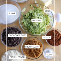 A top-down view of labeled baking ingredients in bowls on a wooden surface, including flour, butter, sugar, eggs, shredded zucchini, chocolate chips, cocoa, brown sugar, protein powder, baking soda, and vanilla.