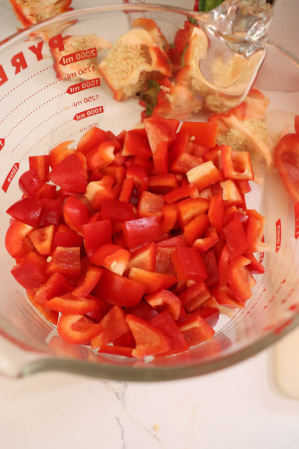 A glass mixing bowl filled with chopped red bell peppers sits on a countertop next to pepper tops and seeds. Measurement markings are visible on the side of the bowl.