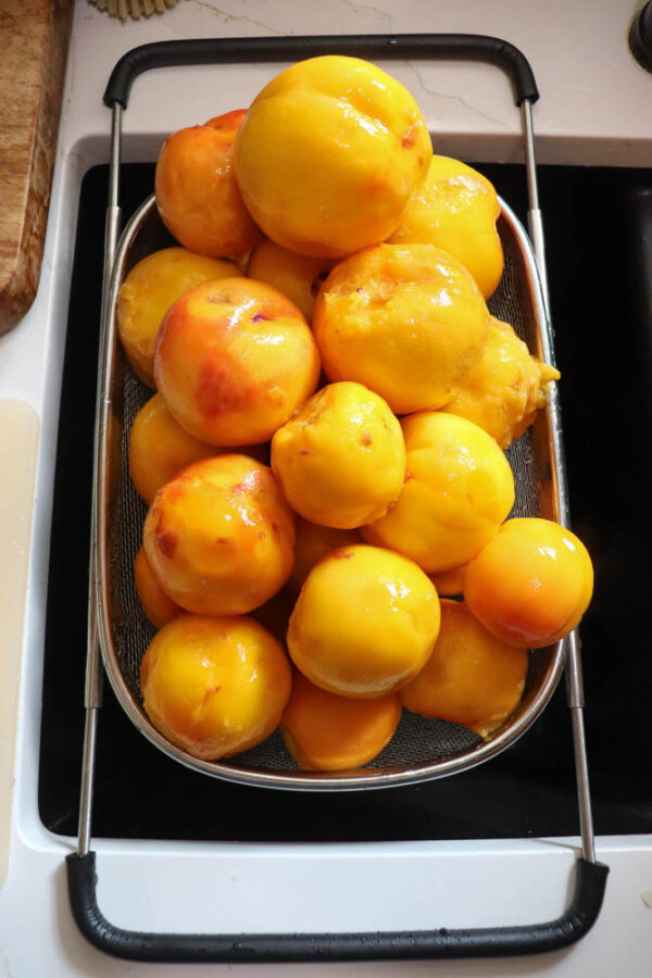 A colander filled with peeled yellow peaches sits over a kitchen sink, with water droplets visible on the fruit.