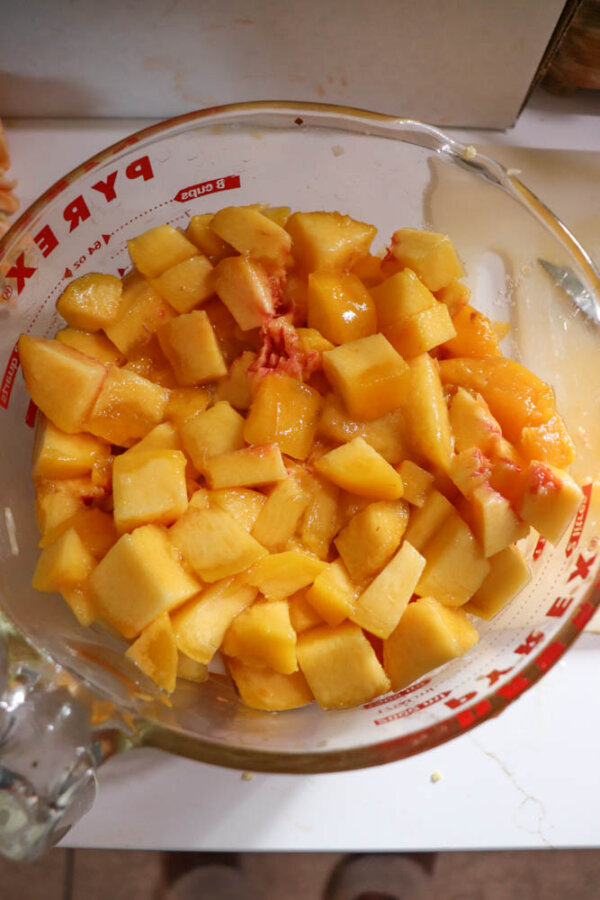 Diced yellow peaches fill a glass Pyrex measuring cup, with some juice pooling at the bottom and peach remnants scattered around the rim.