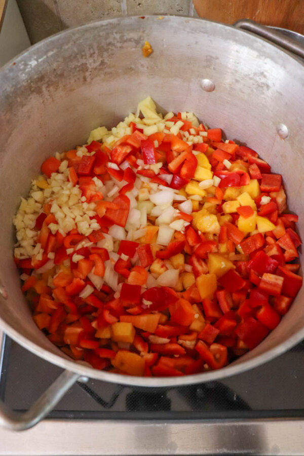 A large metal pot filled with chopped vegetables, including red and yellow bell peppers, onions, and garlic, sits on a stovetop ready for cooking.