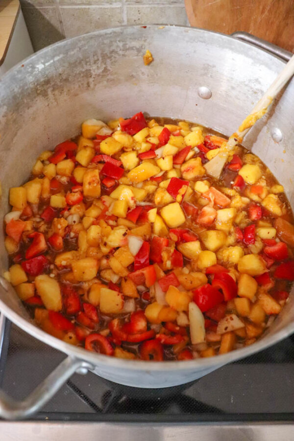 A large pot on a stove filled with chopped apples, red bell peppers, onions, and spices being stirred with a wooden spoon, likely in the process of making chutney or a similar dish.