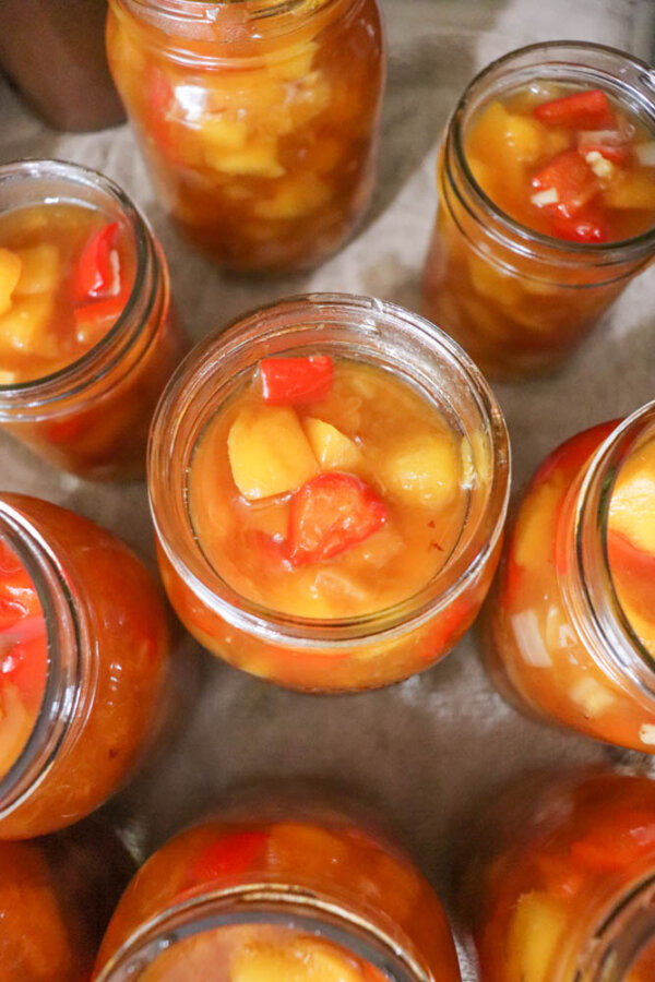 Glass jars filled with chunky peach and red pepper preserves, arranged closely together on a light surface. The contents are colorful and suspended in syrup.