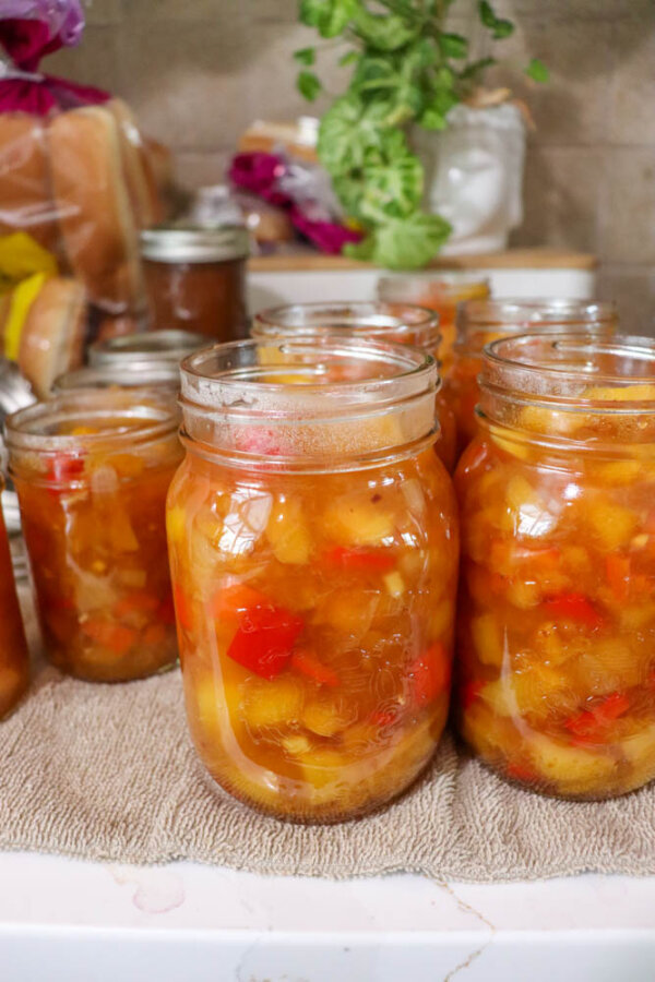 Glass jars filled with homemade chunky peach and red pepper preserves sit on a towel, with more jars and kitchen items blurred in the background.
