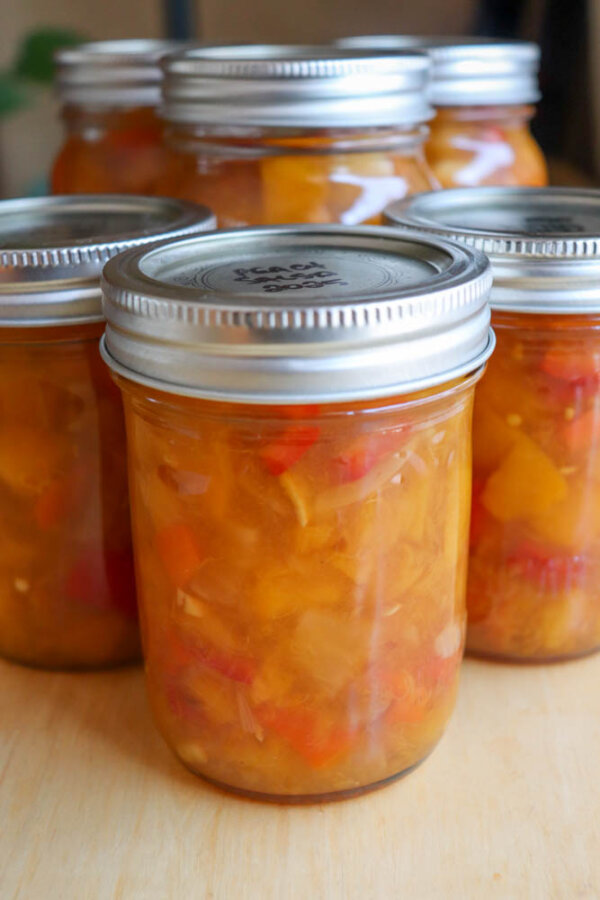 Six glass jars filled with a chunky, orange-yellow relish or preserve with visible pieces of red and yellow vegetables, sealed with silver metal lids, are arranged on a light wooden surface.