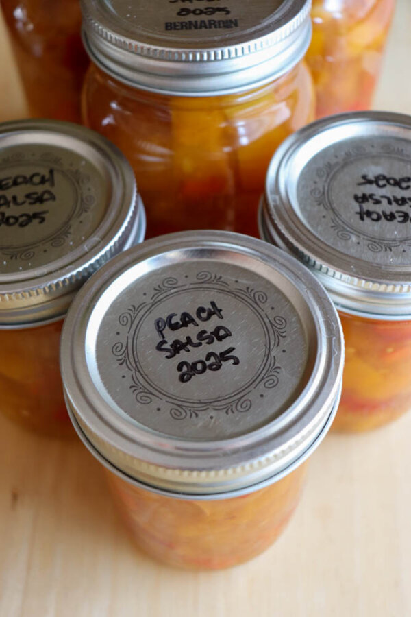 Five jars of homemade peach salsa, labeled “Peach Salsa 2025,” are arranged on a light wooden surface. The jars are sealed with metal lids, and the bright salsa is visible inside.