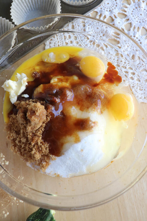 A glass bowl filled with baking ingredients, including eggs, vanilla extract, butter, brown sugar, and granulated sugar, on a wooden surface next to a lace doily and empty cupcake liners.