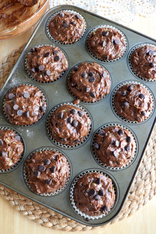 A muffin tin filled with twelve unbaked chocolate chip muffins in paper liners, sitting on a woven placemat, ready to be baked.