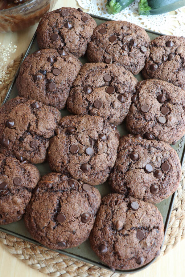A tray filled with freshly baked double chocolate chip cookies, topped with chocolate chips, arranged in three neat rows on a woven placemat.