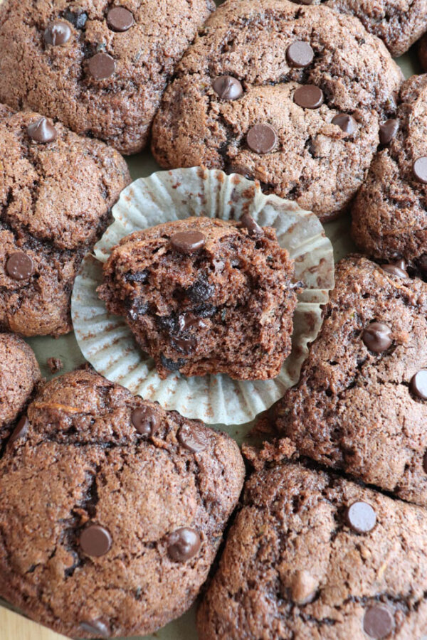 A close-up of several chocolate muffins topped with chocolate chips. One muffin is unwrapped and split open, revealing a moist, chocolatey interior. The muffins are arranged closely together on a flat surface.