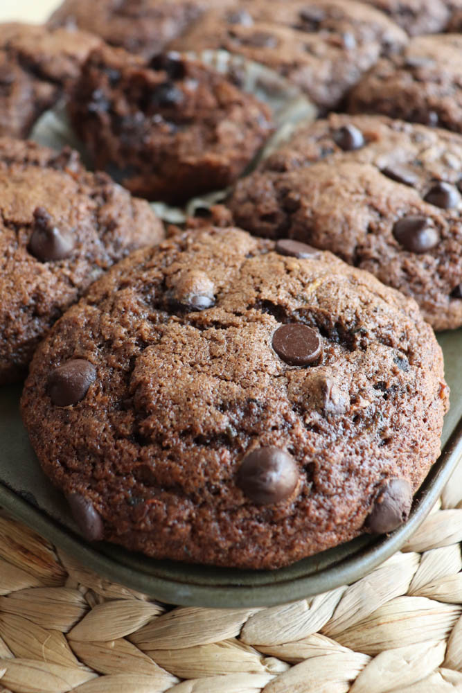 Close-up of a plate filled with large, soft chocolate chip cookies, with several more cookies in the background on a woven placemat.