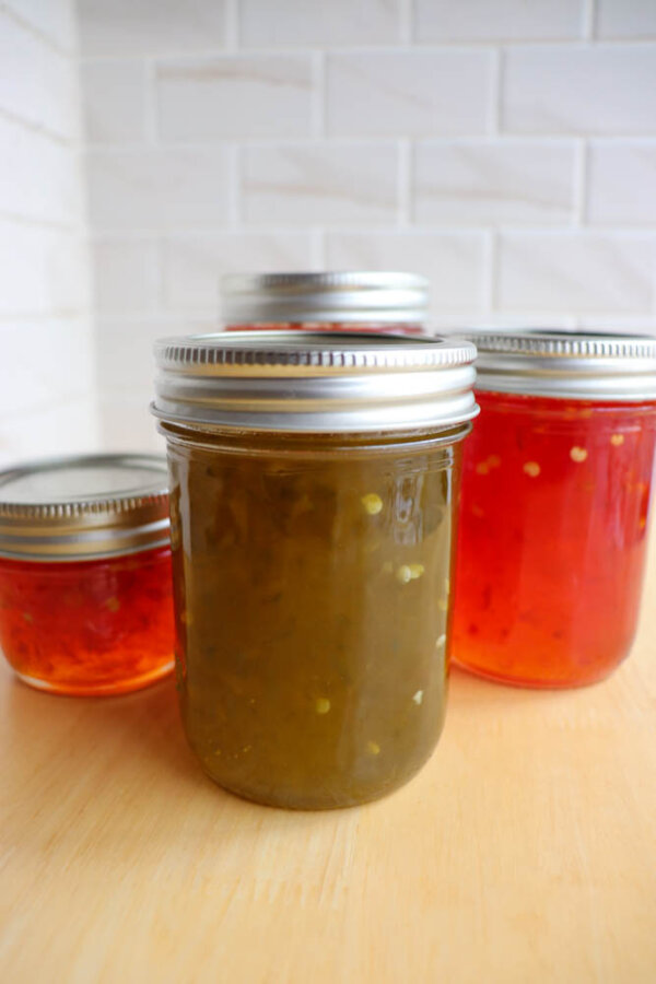 Four glass jars of homemade jelly, two filled with green jelly and two with red jelly, are arranged on a wooden surface in front of a white tiled wall.