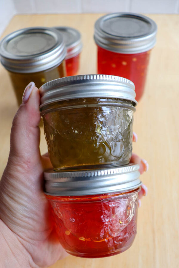 A hand holds two small mason jars of homemade jelly, one yellow-green and one red. Three more jars of jelly sit on a wooden surface in the background.