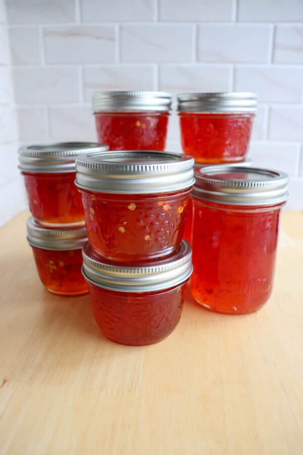 Eight glass jars filled with orange-red jelly, sealed with silver lids, are arranged on a light wooden surface with a white tile background.