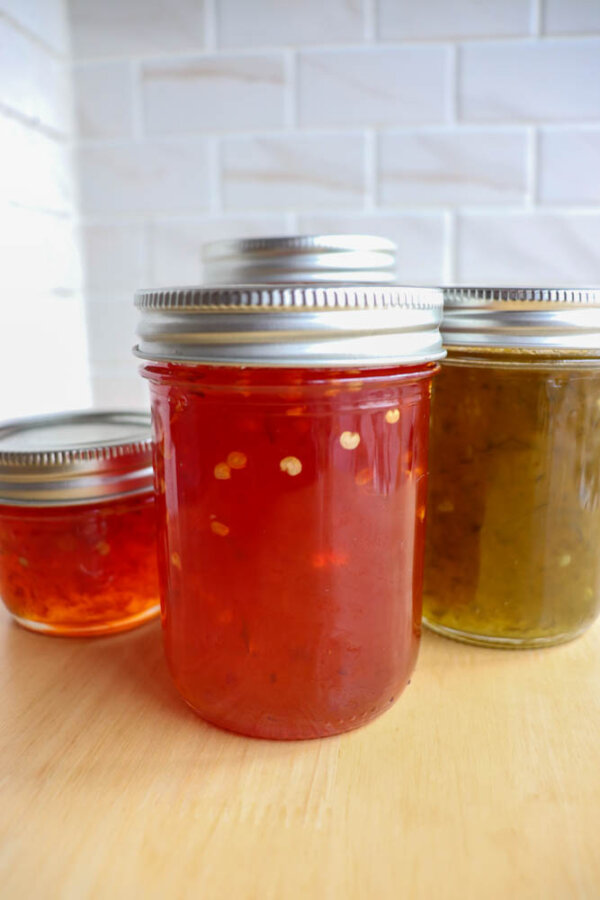 Three glass jars filled with colorful homemade jams or preserves, each with a metal lid, sit on a wooden surface with a white tiled background. The jams are red, orange, and green in color.