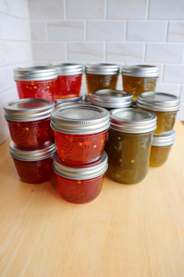 Several small glass jars filled with red and green jelly are arranged on a wooden surface, with metal lids, in front of a white tiled wall.