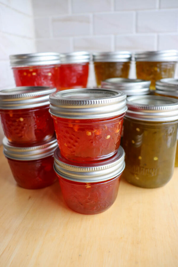 A group of glass jars filled with red and green homemade preserves or jelly, sealed with metal lids, are stacked on a wooden surface with a white tiled background.