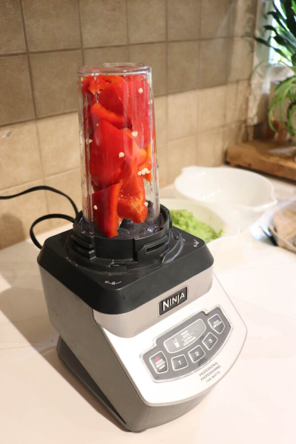 A Ninja blender with a cup filled with chopped red bell peppers sits on a kitchen counter, ready to blend. In the background, a white bowl and some green food are visible.
