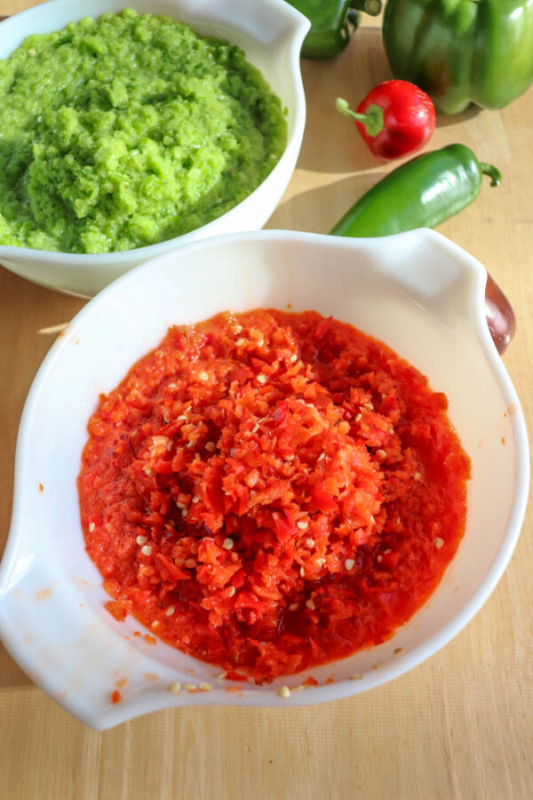 Two white bowls on a wooden surface, one filled with finely chopped green peppers and the other with finely chopped red peppers. Whole green and red peppers are scattered nearby.