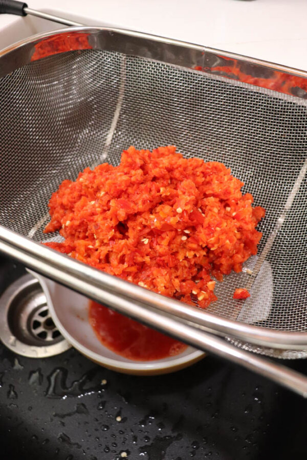 Chopped red peppers are in a metal strainer set over a sink, with liquid draining into a white bowl beneath.