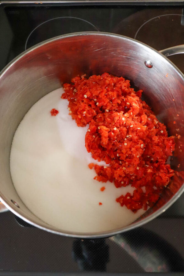 A metal pot on a stovetop contains a pile of granulated white sugar and a mound of finely chopped red chili peppers.