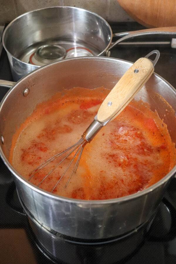 A metal saucepan with a wooden-handled whisk sits on a stove, filled with simmering red sauce. In the background, another pot with jars inside is visible.