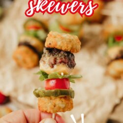 A hand holds a small cheeseburger skewer featuring tater tots, a beef patty, cheese, lettuce, tomato, and pickles. The text reads CHEESEBURGER skewers and SO FUN! above a blurred background.