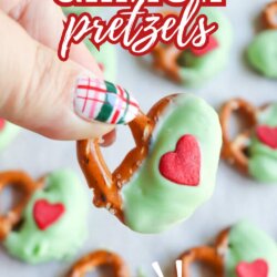 A hand holding a pretzel dipped in green coating with a red heart sprinkle, surrounded by more pretzels on a tray. Text reads: Grinch pretzels, SO FUN, homemadeheather.com.