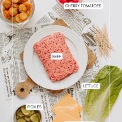 Flat lay of ingredients for a dish: a plate with raw ground beef, a bowl of tater tots, cherry tomatoes, pickles, sliced cheese, and lettuce leaves on a table with newspaper and toothpicks.