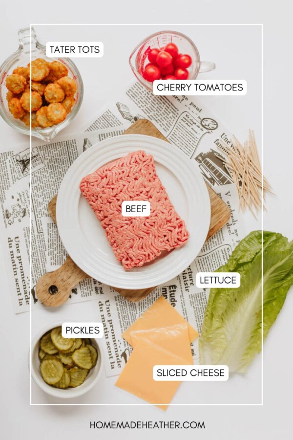Flat lay of ingredients for a dish: a plate with raw ground beef, a bowl of tater tots, cherry tomatoes, pickles, sliced cheese, and lettuce leaves on a table with newspaper and toothpicks.