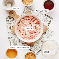 Top-down view of small bowls containing chopped bacon, brown sugar, maple syrup, diced onion, minced garlic, apple cider vinegar, and spices, all labeled, on a white background with a printed mat.