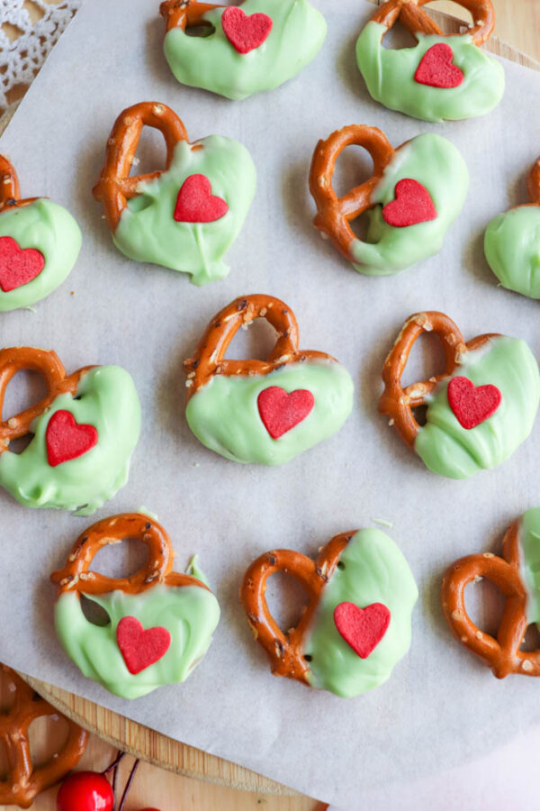 Pretzels partially dipped in green coating are arranged on parchment paper. Each pretzel is decorated with a small red heart-shaped candy on the green-coated section.