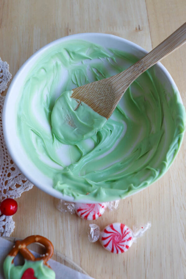 A white bowl with green icing remnants and a wooden spoon inside. Two peppermint candies and a decorated pretzel are nearby on a light wooden surface.