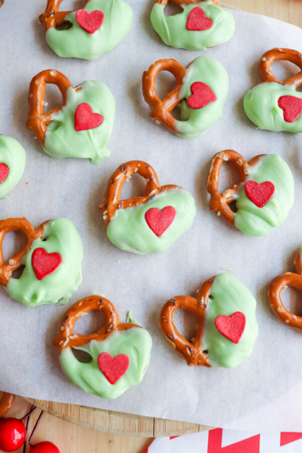 Pretzels partially dipped in green coating, each decorated with a red heart-shaped candy, are arranged on parchment paper. The treats have a festive, playful appearance, perfect for a themed celebration.