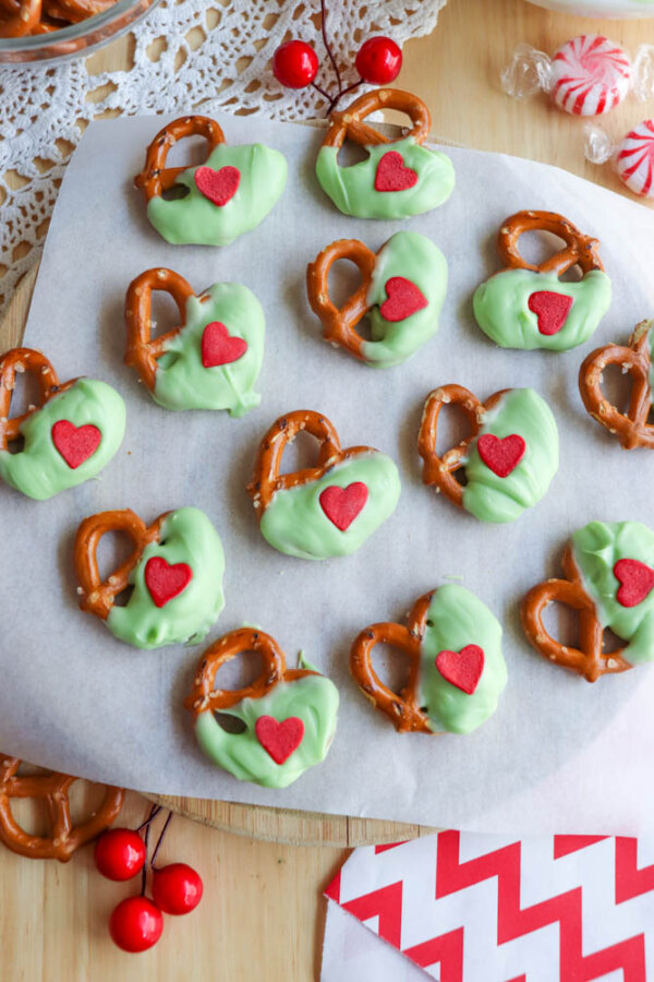 Pretzel twists half-dipped in green chocolate, each topped with a small red heart candy, are arranged on parchment paper atop a wooden surface. Peppermint candies and red decorations are seen nearby.
