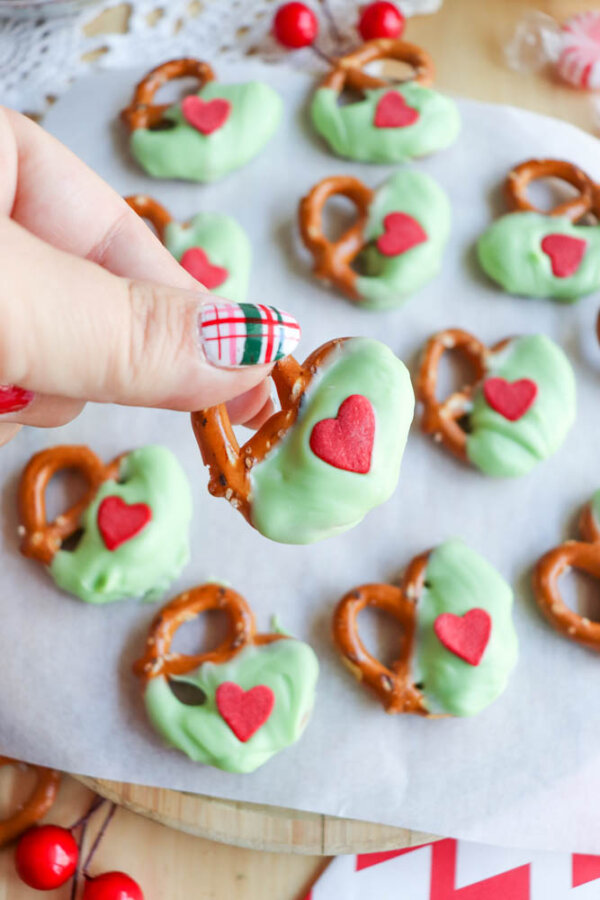 A hand with plaid nail art holds a pretzel half-dipped in green coating, decorated with a red heart. More similarly decorated pretzels are arranged on parchment paper in the background.