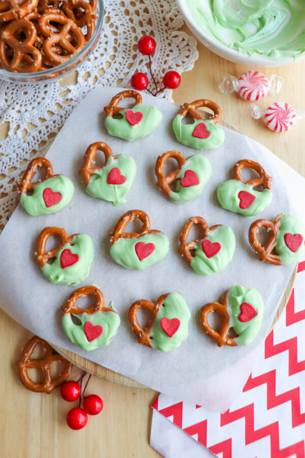 A tray of mini pretzels half-dipped in green coating, each topped with a red heart sprinkle, sits on parchment paper with peppermint candies, red cherries, and a bowl of green coating nearby.