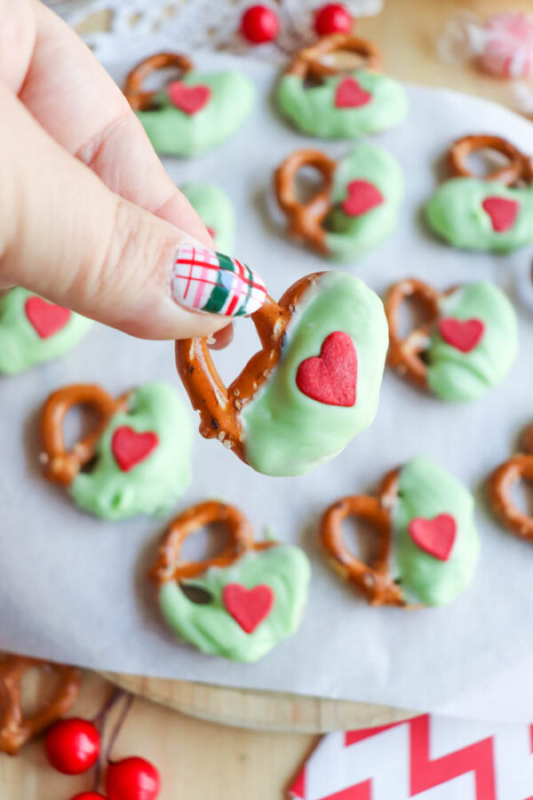 A hand with plaid-patterned nails holds a pretzel dipped in green coating and decorated with a red heart. More pretzels with the same decoration are on parchment paper in the background.