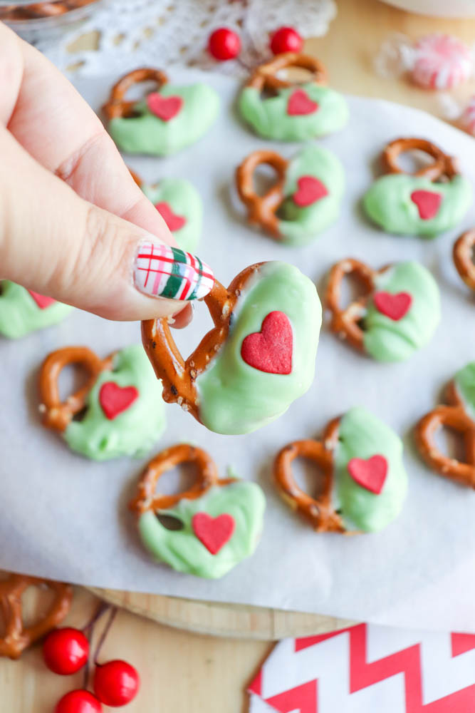 A hand with festive plaid nail art holds a pretzel half dipped in green frosting and decorated with a red heart. More green-frosted pretzels with red hearts are on parchment paper in the background.