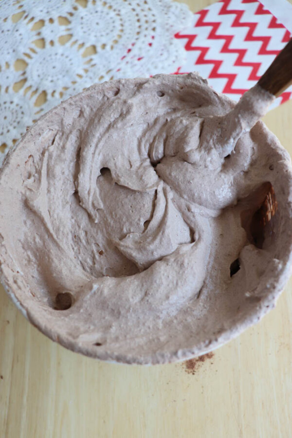 A large bowl filled with fluffy chocolate mousse being mixed with a wooden spoon, sitting on a light wooden surface near a white lace doily and red chevron-patterned paper.