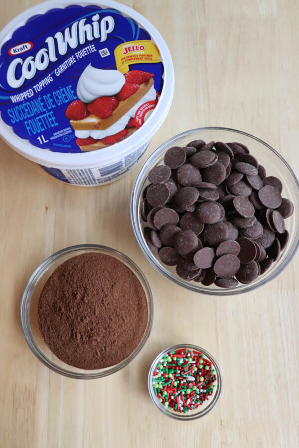 Four ingredients on a wooden surface: a tub of Cool Whip, a bowl of chocolate melting wafers, a bowl of cocoa powder, and a small bowl of red, green, and white holiday sprinkles.