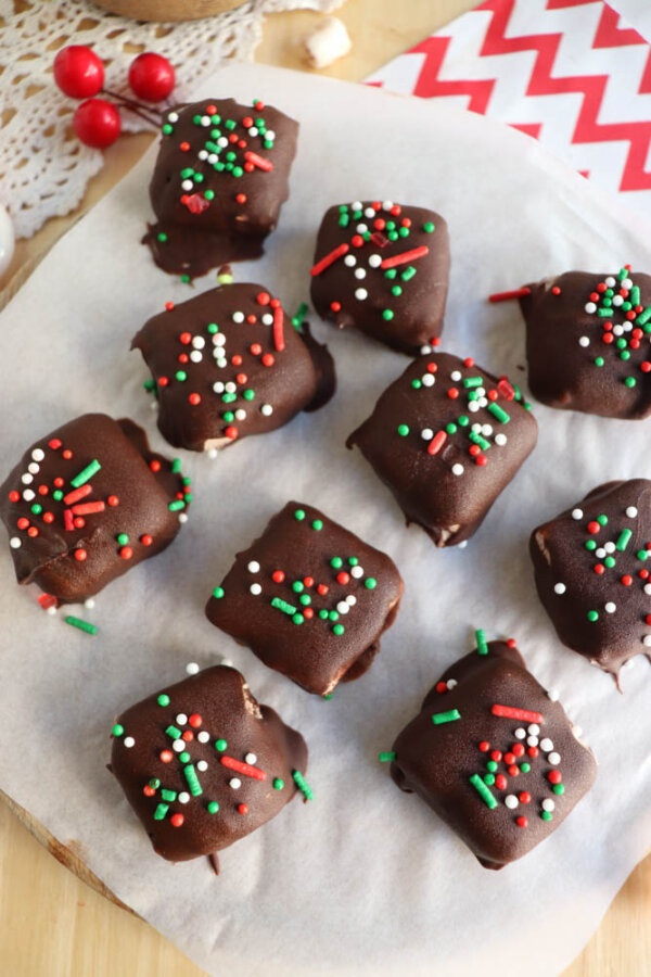 Chocolate-covered square treats topped with red, white, and green sprinkles are arranged on parchment paper. A few red and white candies and a red-and-white chevron napkin are visible nearby.
